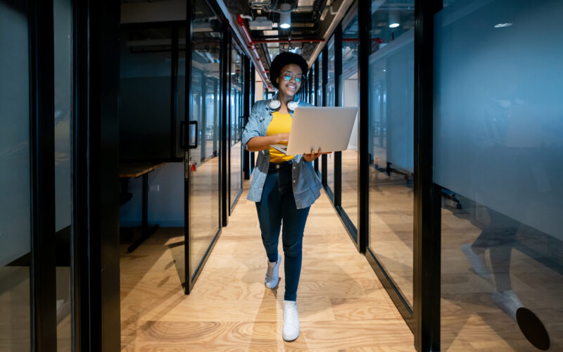 Woman walking through a glass-walled office corridor carrying a laptop
